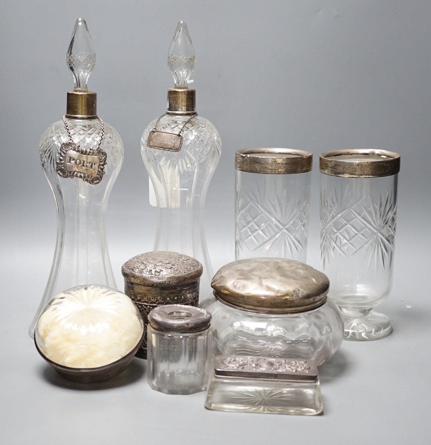 A collection of silver mounted glass items, including a pair of waisted decanters with two silver wine labels, a pair of vases, powder jar etc. and an Indian white metal jar and cover.